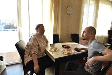 Man and woman having a casual breakfast at a dining table in a bright room with sunlight coming through the large window. Plates with simple food and a cup of coffee create a relaxed morning scene.