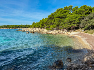 Obraz premium Scenic coastline showcasing a pristine pebbled beach bordering crystal clear turquoise water and lush green pine trees, under a vibrant blue sky on the sunny shores of Rab Island, Croatia.