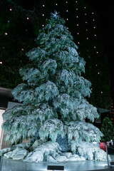 A festive Christmas tree display featuring sparkling ornaments, twinkling lights, and seasonal decorations. The tree is adorned with baubles, ribbons, garlands, and a bright star on top, creating a wa
