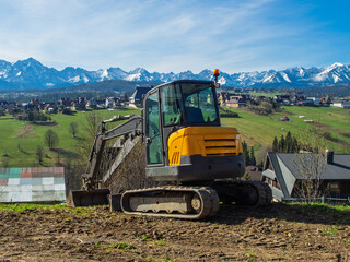 A small yellow tracked excavator works on a construction site, moving soil on a sunny day with the picturesque Polish Tatra Mountains and traditional rural houses in the background.