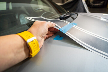 Worker installing car decals in garage, close-up of hand holding black vinyl film. Car decal application on hood.
