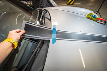 Worker installing car decals in garage, close-up of hand holding black vinyl film. Car decal application on hood.