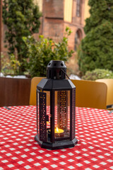 Black metal lantern with lit candle on red and white checkered tablecloth in outdoor cafe setting...