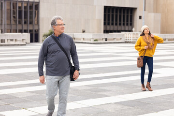 Senior couple wearing athletic attire and mustard-yellow sweater walking across plaza carrying bags