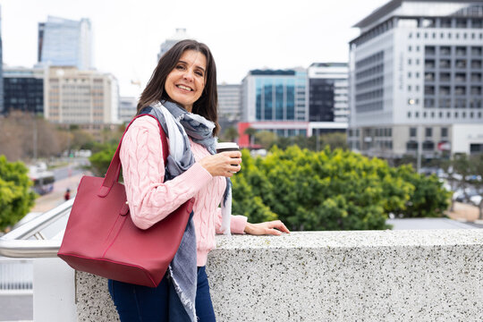 Mature adult woman leaning against railing holding coffee cup on city overlook, copy space