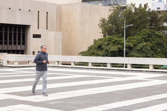 Senior man jogging across tiled urban plaza wearing gray activewear past balustrade near lamppost