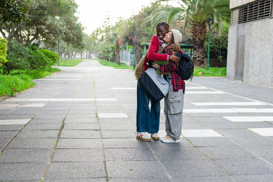 Diverse female friends hugging on urban campus walkway, carrying tote bag and backpacks, copy space