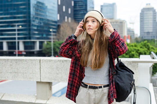 Woman adjusting white over-ear headphones while standing on elevated concrete terrace with backpack