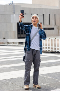 Asian male smiling and holding smartphone and coffee cup on rooftop terrace with messenger bag