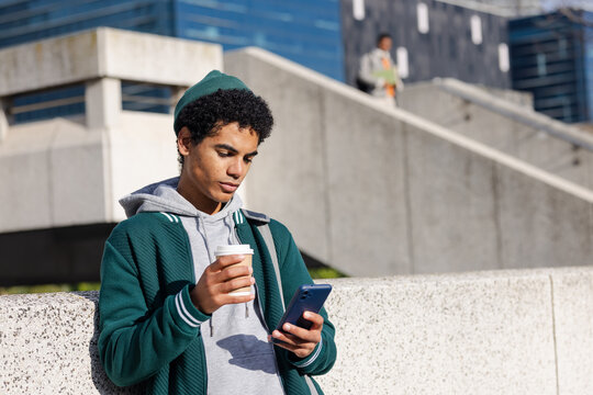 African American student leaning on wall checking smartphone, sipping coffee at plaza, copy space