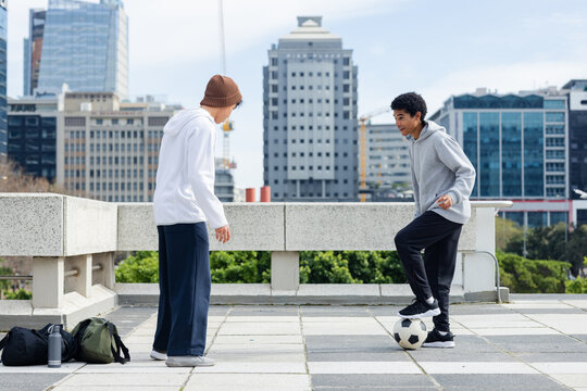 Diverse male friends playing soccer on rooftop terrace by railing with backpacks and water bottle