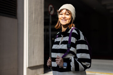 30s woman standing on street by no-parking sign wearing beanie, backpack and smartwatch, copy space