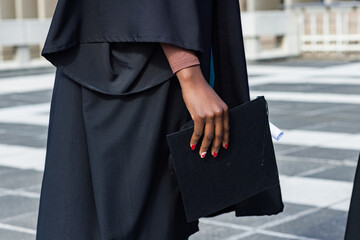 Graduate standing on patterned courtyard holding mortarboard and diploma, building facade blurred