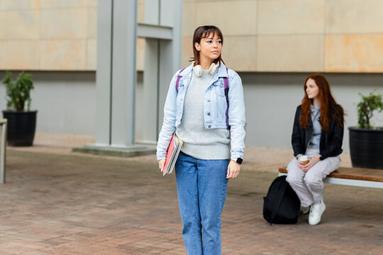 Classmates walking across courtyard carrying notebooks and wearing headphones near friend on bench
