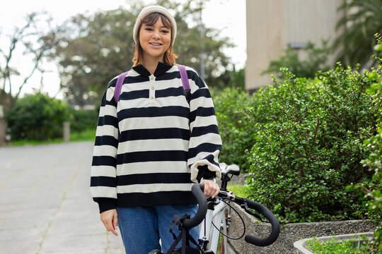 Woman standing on paved walkway holding road bicycle handlebars and carrying purple backpack
