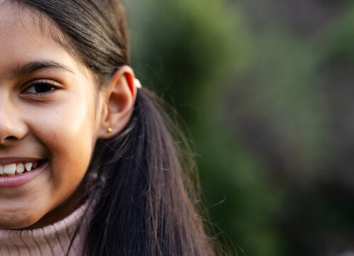Indian girl smiling showing cheek and eye in garden wearing pink turtleneck sweater copy space