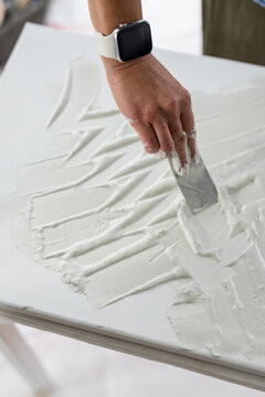 Mature adult woman smoothing white paste on rectangular panel at workshop table using metal spatula