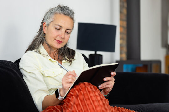Senior woman sitting on sofa at home writing in notebook beside brick fireplace, copy space