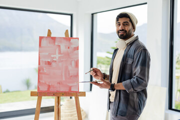 Indian man standing beside easel holding brush and palette wearing headphones in studio, copy space