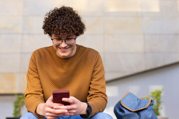 Man sitting on bench at urban plaza holding smartphone and checking messages next to backpack