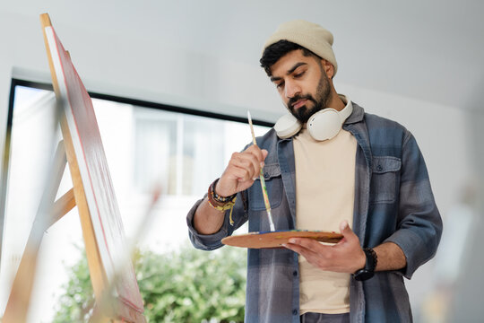 Indian male painter mixing colors on canvas at art studio with palette, brush, headphones - Powered by Adobe