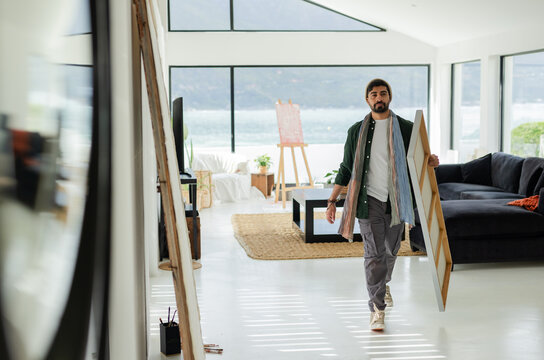 Indian man walking at open-plan living area doubling as art studio carrying stretcher-framed canvas
