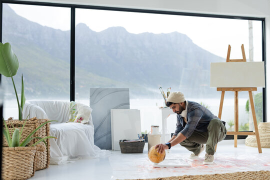 Indian man crouching on plastic sheet in living room, applying paint to floor canvas near easel