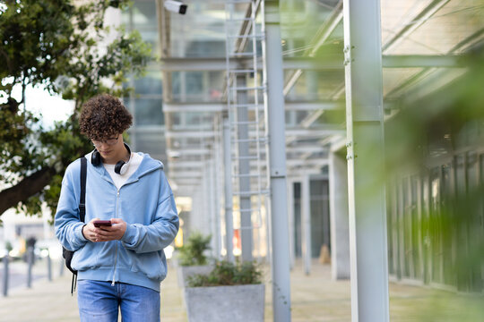 Man walking on covered walkway wearing hoodie and headphones while checking smartphone, copy space