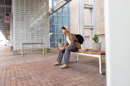 Adult Asian man wearing beanie sitting on transit bench checking phone with backpack and signage