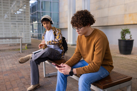 Diverse male friends sitting on plaza bench checking wristwatch, scrolling smartphone with notebook