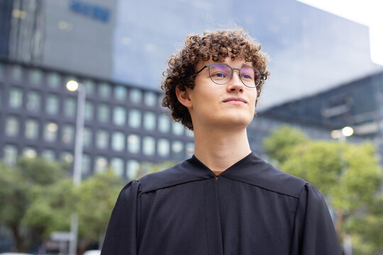Male graduate looking up wearing black robe and round eyeglasses outside glass building, copy space
