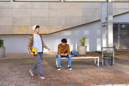 Diverse male students walking with yellow notebook while scrolling smartphone on bench in courtyard