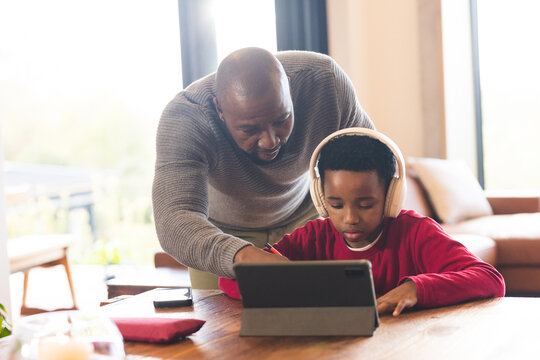 African American father and son working using tablet and stylus with headphones in home living room - Powered by Adobe