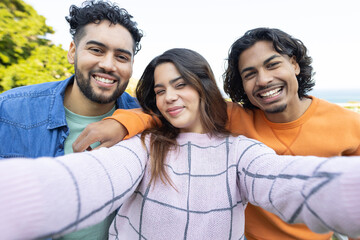 Friends leaning in and capturing selfie at coastal park with smartphone under sunny sky