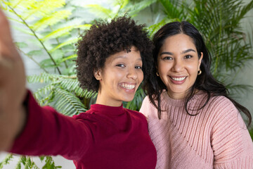 Female friends standing in plant-filled room holding smartphone and taking selfie amid lush ferns