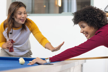 Female friends lining up shot on blue-felt pool table in game room holding red drink bottle