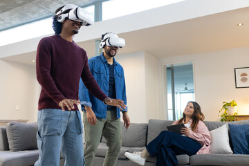 Diverse friends wearing white VR, reaching out near sofa while woman holding tablet watching