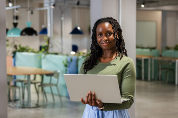 Woman holding laptop at waist, wearing green top and light blue skirt in coworking space