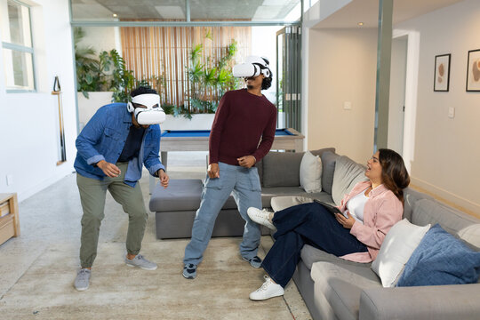 Diverse friends wearing white VR headsets interacting in modern game room with tablet, pool table - Powered by Adobe