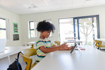 African American child sitting at white table in bright classroom touching tablet on stand