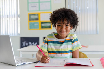 African male youth student sitting at class desk writing in notebook with pink pencil, striped tee