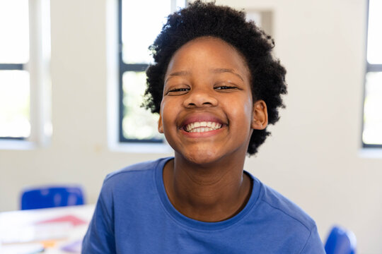 Adolescent African female wearing blue shirt smiling and leaning in classroom near windows, papers