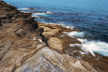 Dynamic high angle view of layered, eroded rock strata meeting the deep blue ocean. White waves crash against the rugged shoreline