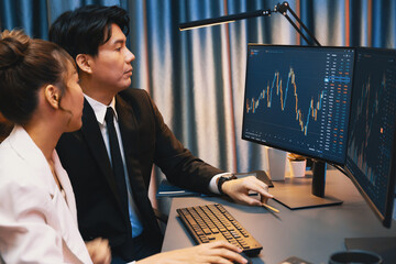 Smiling Asian business traders pointing to dynamic stock exchange data screen profit value online website program at night time at home office. Analyzing currency rate value on data report. Infobahn.