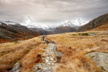 Hiker Climbs Hill Through Dry Grassland While The Matterhorn Rises To The Cloudy Sky