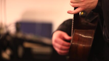 A male guitarist plays an acoustic wooden guitar under warm concert lighting. Close-up shows his large hands strumming, the classical body shape, and the intimate mood of live performance.