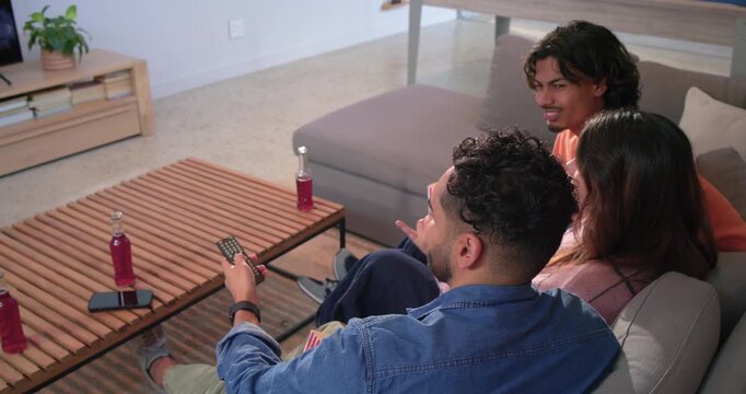 Diverse friends pressing remote and navigating menu on sofa, offering popcorn and selecting show