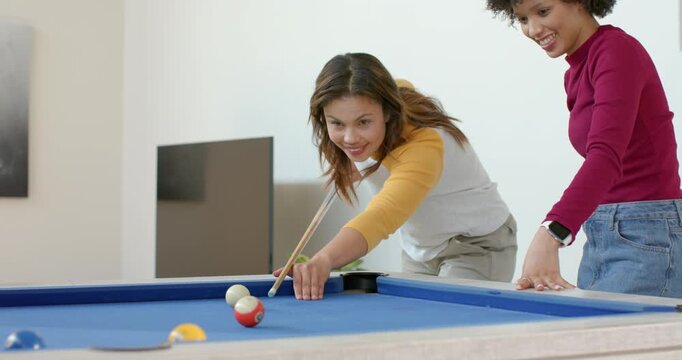Two female friends playing pool on blue-felt table at home, cue ball striking ball high-fiving