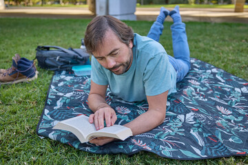 Middle aged man lying on a patterned picnic blanket and reading a book in a sunny green park.