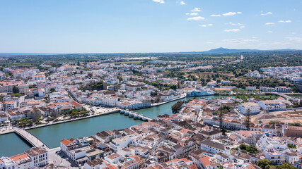 Tavira in Algarve is captured from above, showing its buildings, Gilao river, and green areas under a clear sky with some clouds